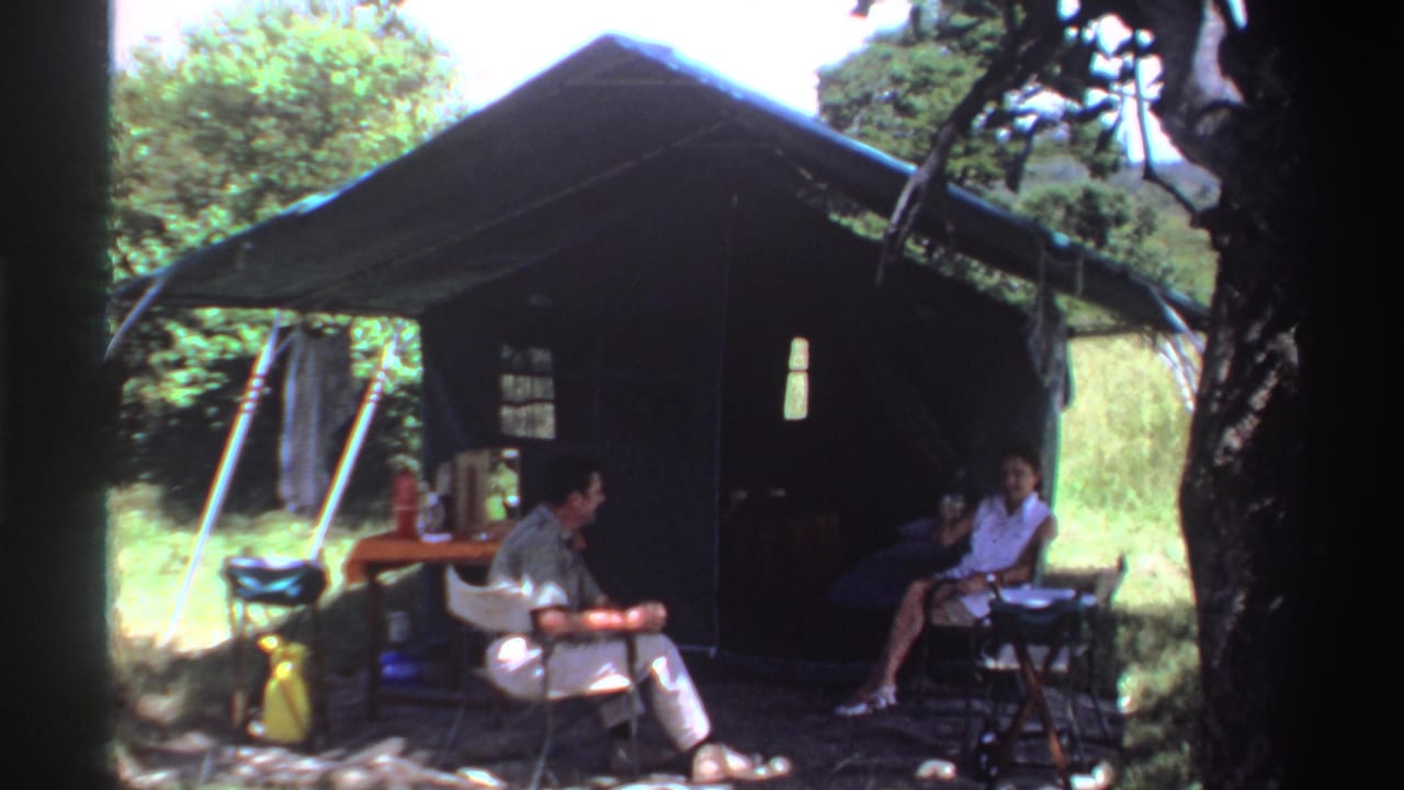 Two adults sit under a green tent with a small table in a wooded outdoor setting.