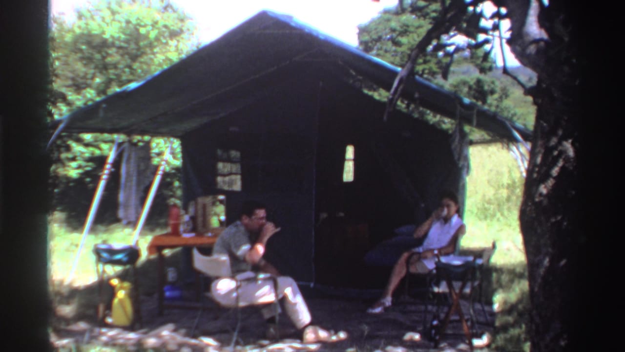 In Nigeria, 1969, two adults sit under a canvas tent as one brings a cup or glass toward his face.