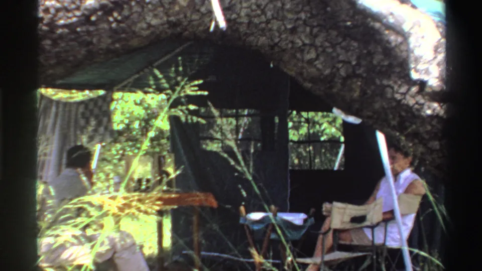 Closer 1969 Nigeria 8mm view of adults resting in chairs below a fallen tree trunk, with fabric shade and vegetation nearby.