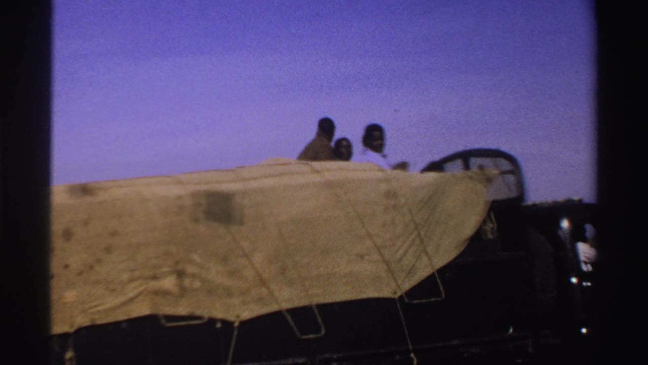 The truck continues past in rural Nigeria, with seated riders visible above the beige covering and open fields behind it.