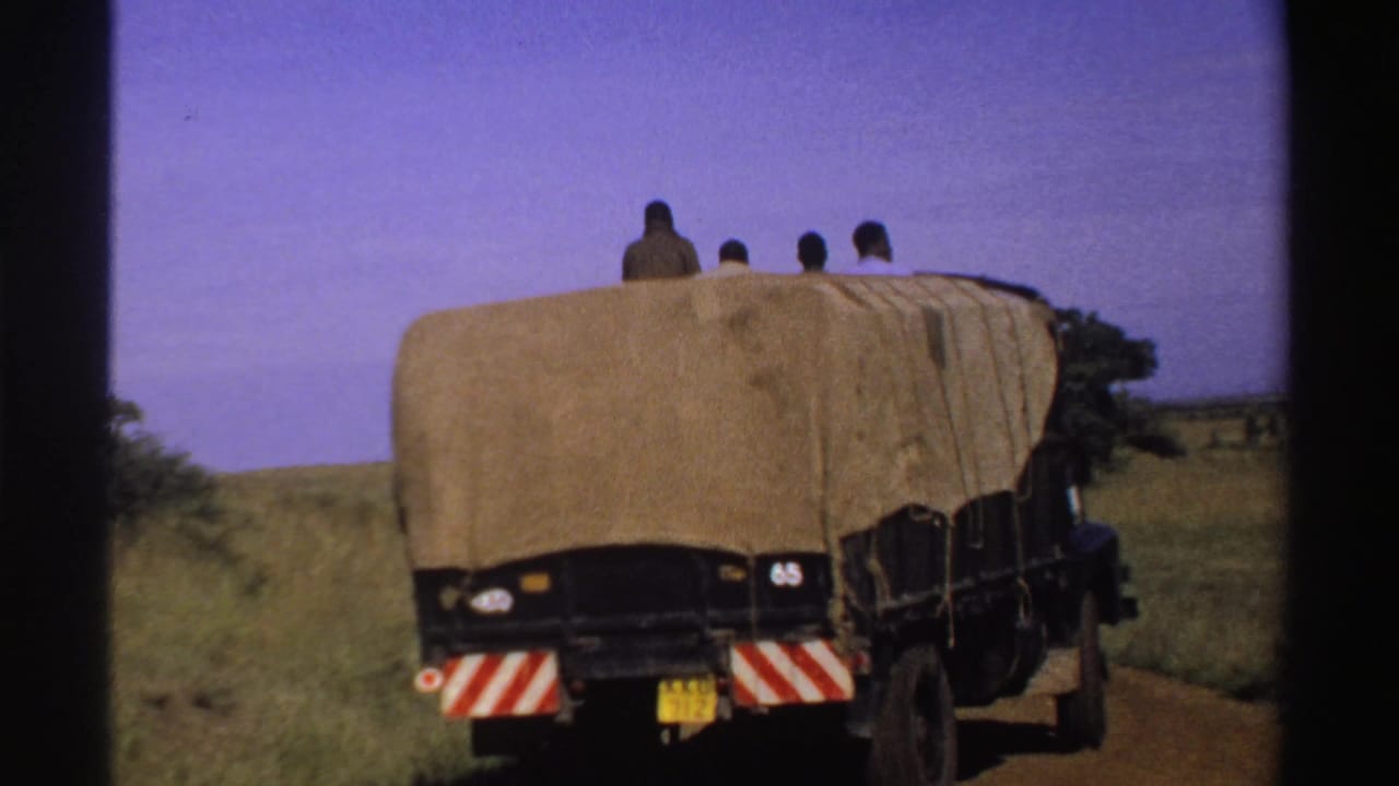 A covered truck drives away along a dirt road in Nigeria, with several riders still visible above the rear of the tarp.