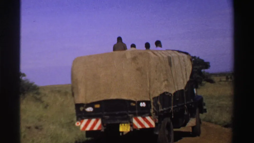 A covered truck drives away along a dirt road in Nigeria, with several riders still visible above the rear of the tarp.