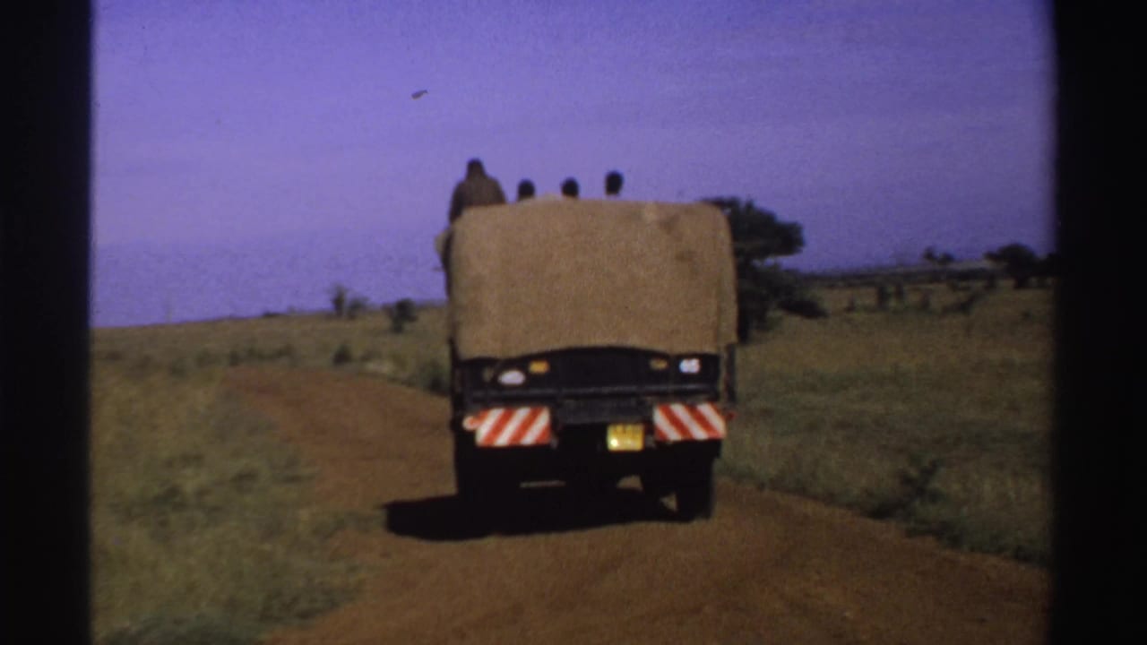 The truck moves farther away on the dirt road, framed by grassy countryside and sparse trees under a purple-blue sky.