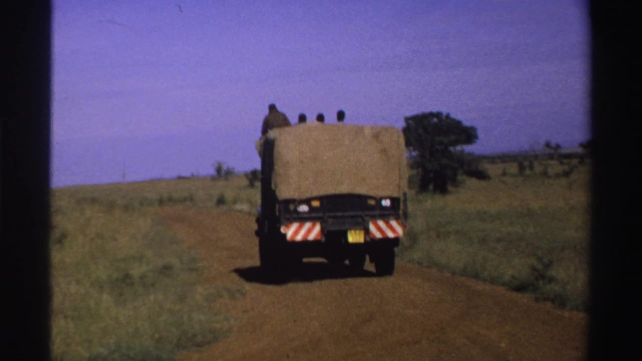 1969 Nigeria 8mm frame showing a tan open-backed truck departing along a reddish dirt road with riders silhouetted in the rear.