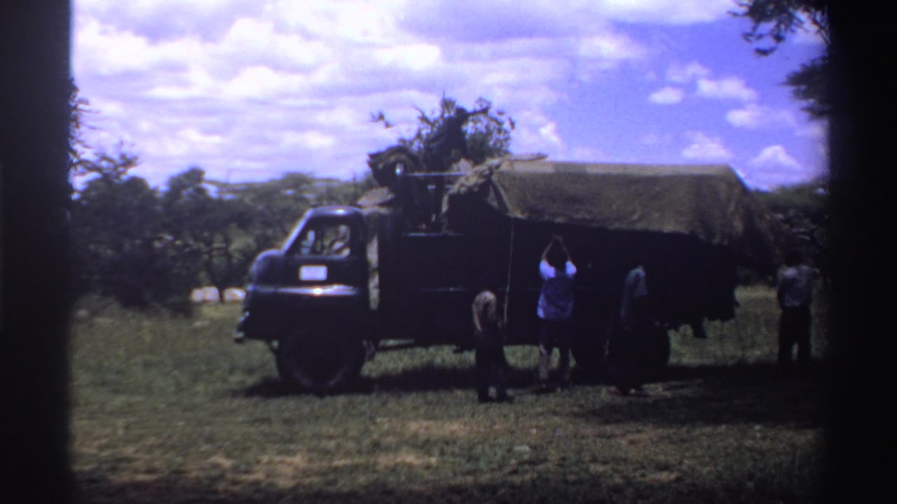 1969 Nigeria 8mm frame showing several people beside a dark canvas-covered truck in a grassy field with trees and clouds behind them.