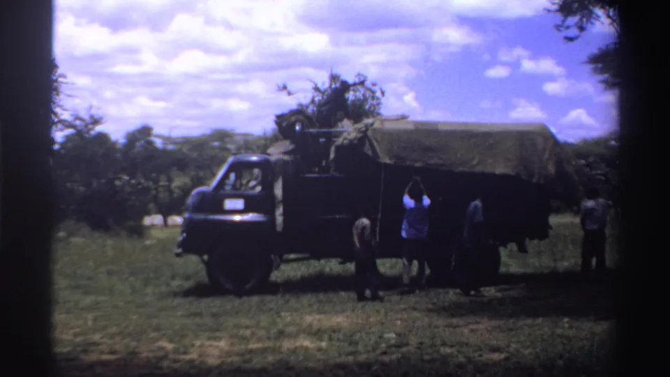 1969 Nigeria 8mm frame showing several people beside a dark canvas-covered truck in a grassy field with trees and clouds behind them.