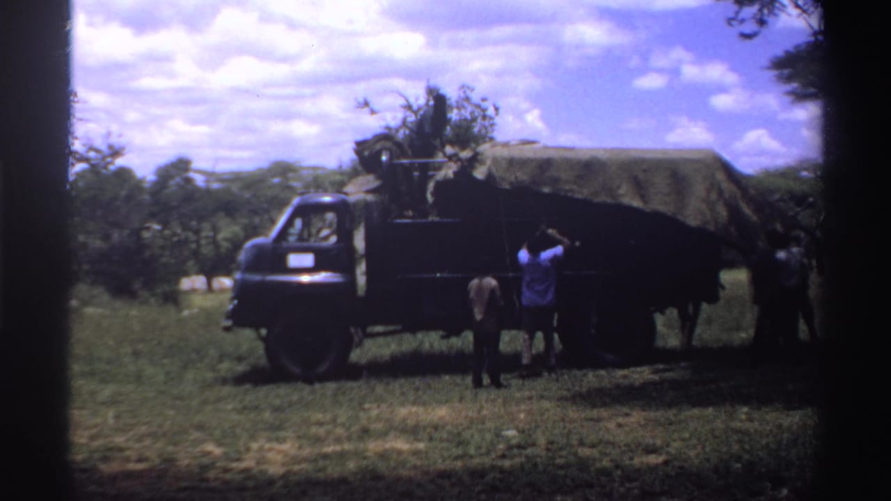 1969 Nigeria home-movie frame with people near a canvas-covered truck, one person reaching toward the tarp while others stand nearby.