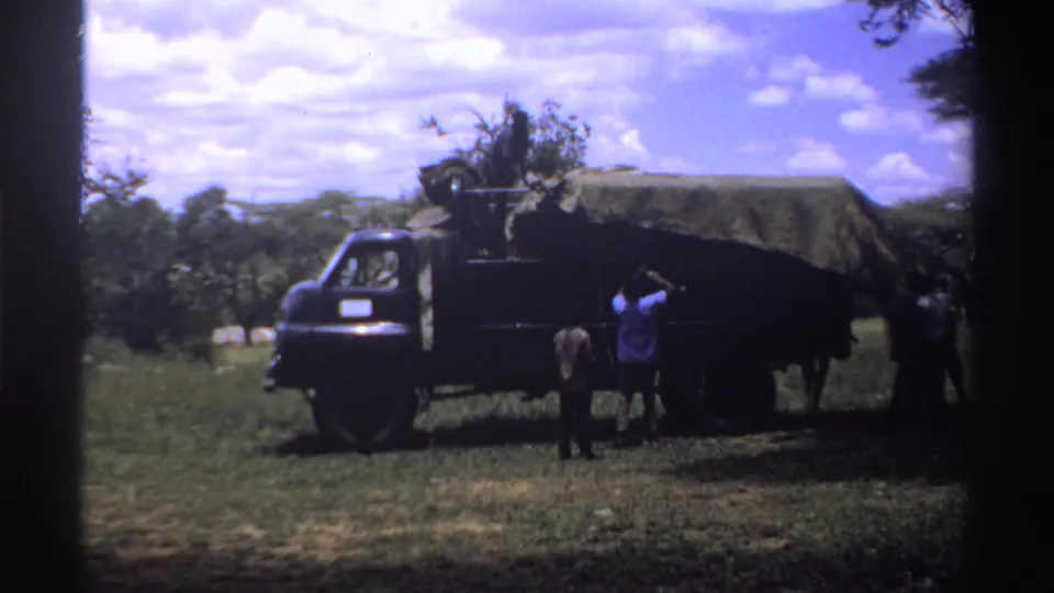 1969 Nigeria home-movie frame with people near a canvas-covered truck, one person reaching toward the tarp while others stand nearby.