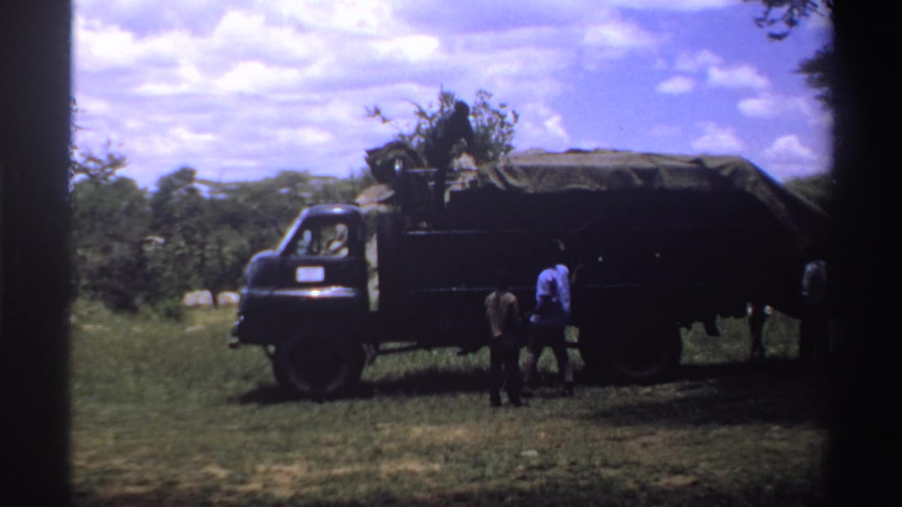 1969 Nigeria 8mm frame showing the side of a dark truck with a covered load and people standing close to the cargo area in a field.