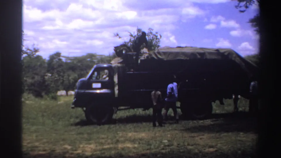 1969 Nigeria 8mm frame showing the side of a dark truck with a covered load and people standing close to the cargo area in a field.