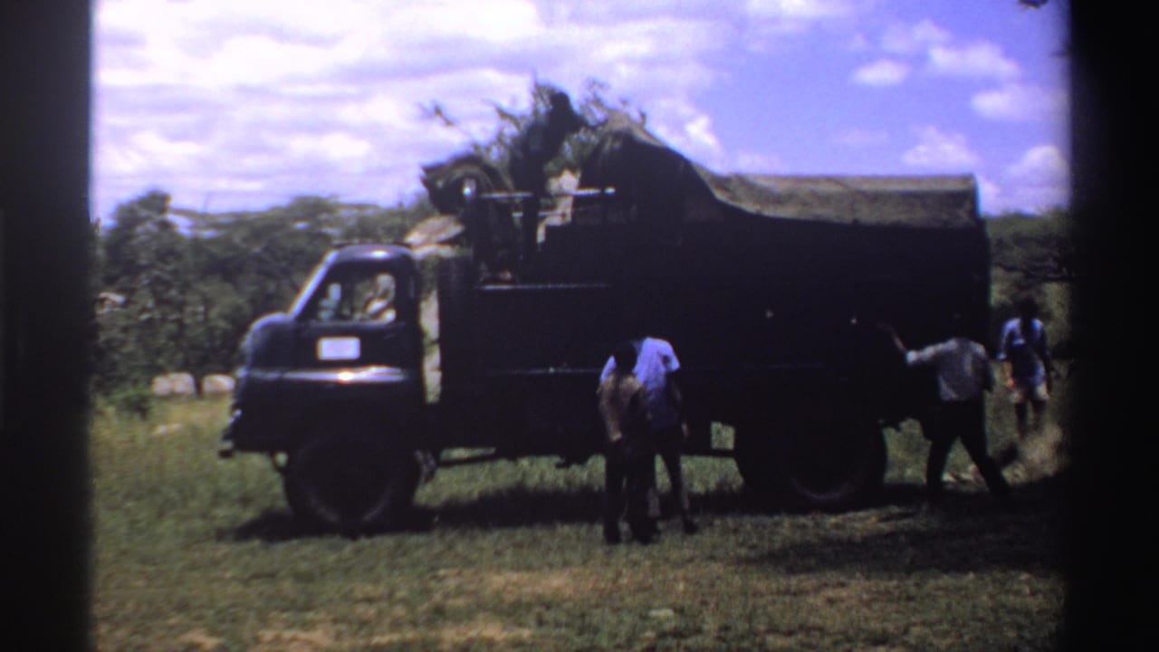 1969 Nigeria home-movie frame showing people unloading or adjusting material near a dark canvas-covered truck in a grassy field.