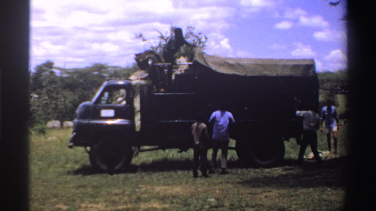 Men gather beside a dark tarp-covered truck in grassy bushland.