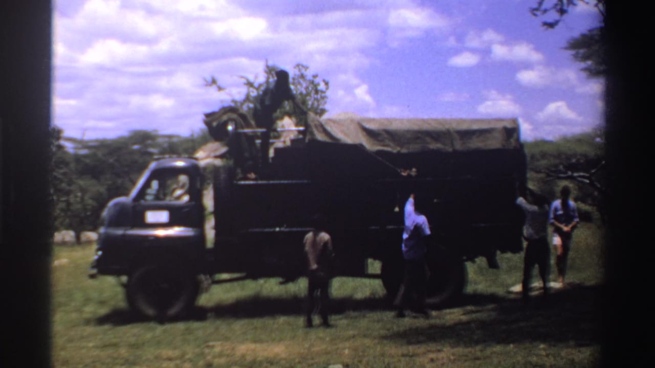 1969 Nigeria frame with men positioned around a dark military-style truck in grassy, tree-dotted bushland.
