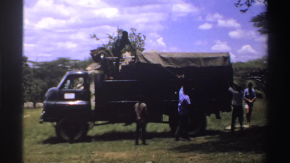 1969 Nigeria frame with men positioned around a dark military-style truck in grassy, tree-dotted bushland.