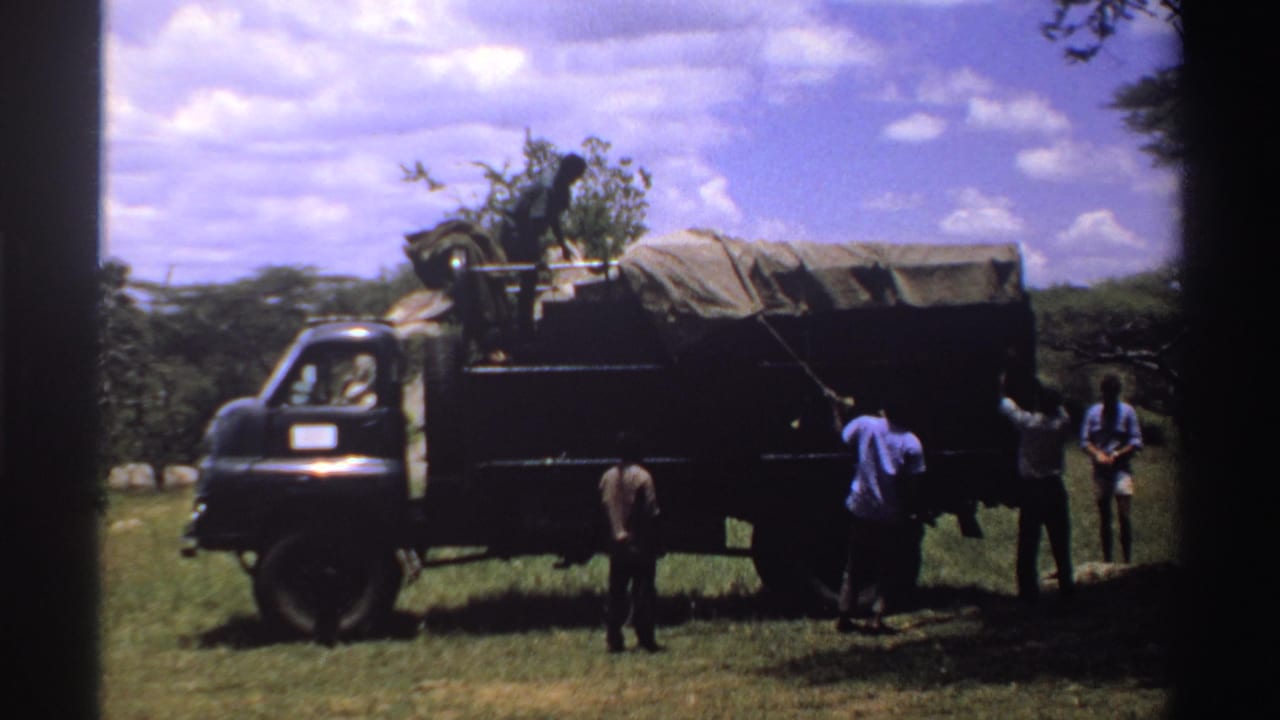 1969 Nigeria 8mm frame showing men at the side of a dark cargo truck, with one figure bent near the vehicle.