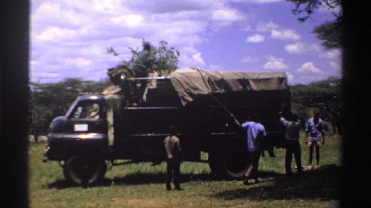 1969 Nigeria frame showing a man extending an arm toward the tarp-covered side of a dark truck as others stand nearby.
