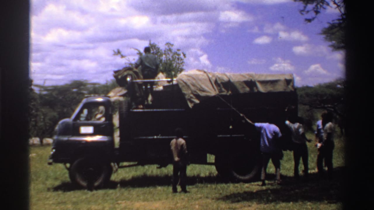 Men stand beside a canvas-covered truck in a grassy field under a clouded sky.