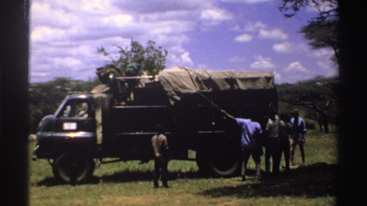 Men in 1969 Nigeria appear to work around a tarp-covered truck parked in grass beneath a bright sky.
