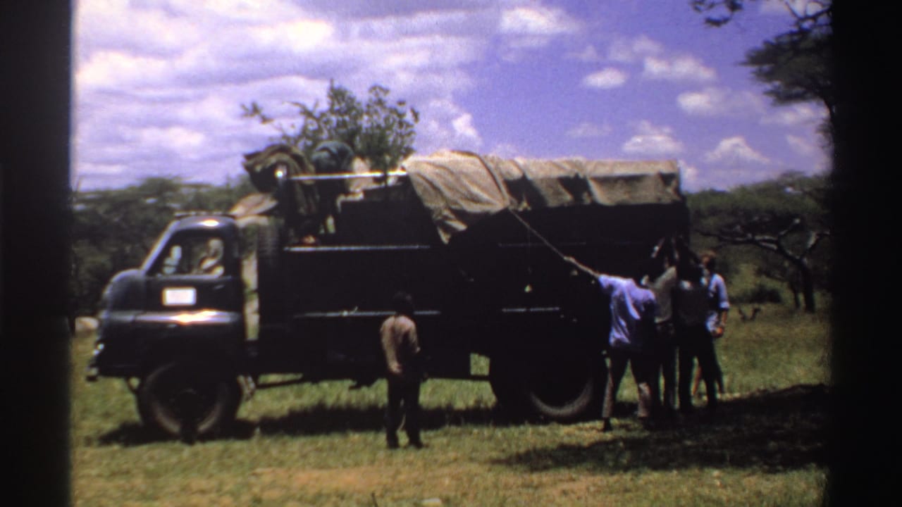 Home-movie frame from Nigeria in 1969 showing men gathered by a dark truck with a covered load in a rural field.