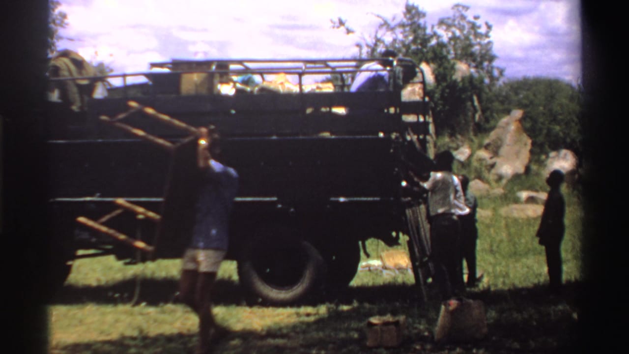1969 Nigeria frame showing a man walking past a loaded truck as others handle items near rocks and grass.