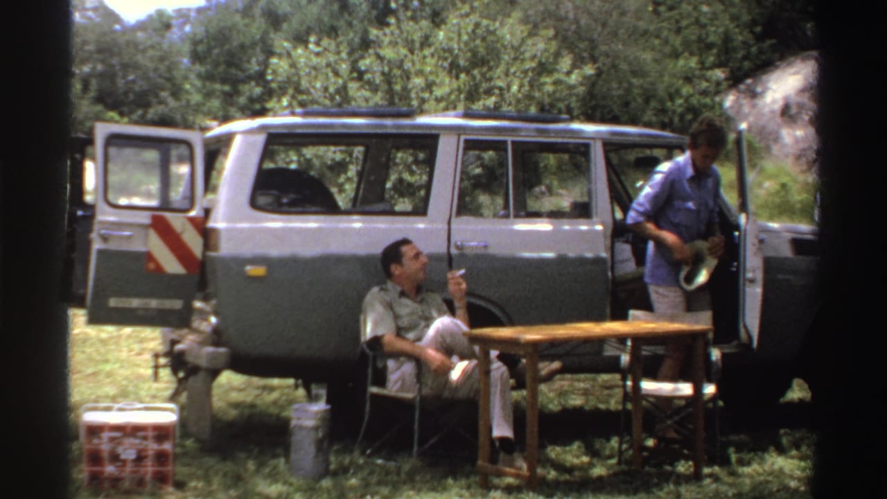 Two adult men beside a two-tone SUV, with one seated smoking near a small table.