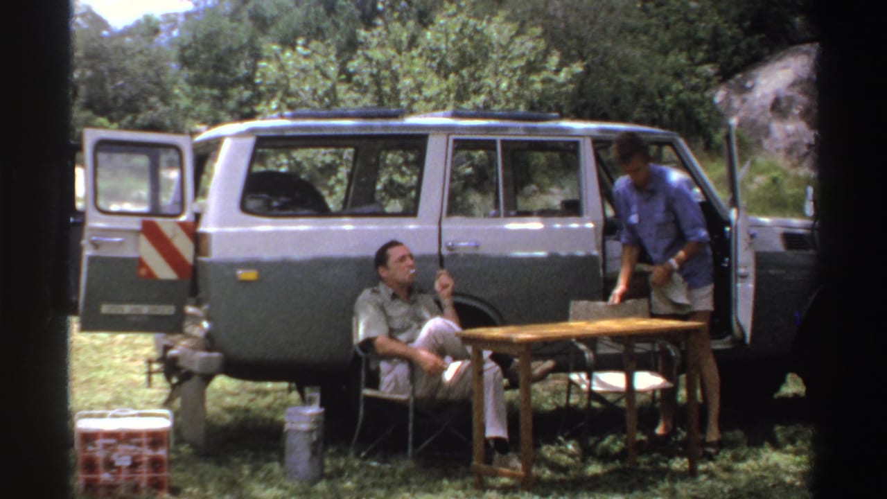 In this Nigeria 1969 roadside frame, a seated man smokes while a man in a blue shirt stands beside the open vehicle door.