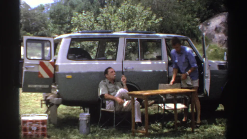 In this Nigeria 1969 roadside frame, a seated man smokes while a man in a blue shirt stands beside the open vehicle door.