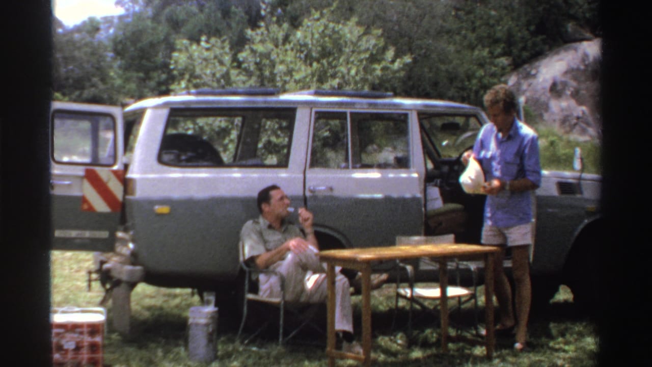 1969 Nigeria 8mm frame showing the standing man holding a hat beside a small table while another man sits smoking by the SUV.