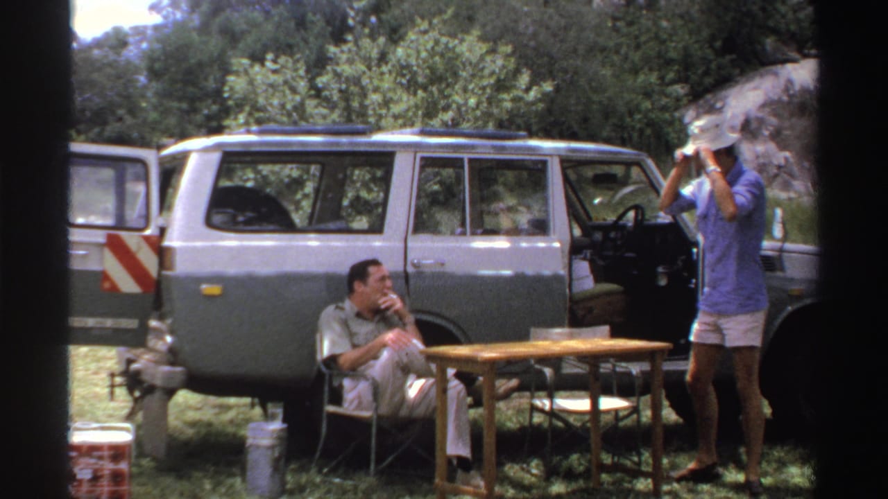 In this late-1960s Nigeria frame, the standing man raises a light hat beside the open SUV while the seated man smokes nearby.