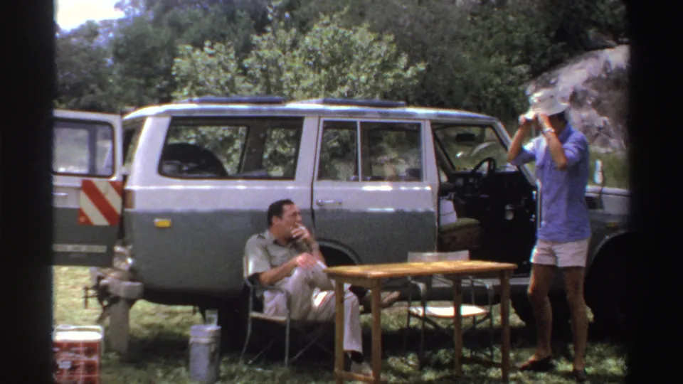 In this late-1960s Nigeria frame, the standing man raises a light hat beside the open SUV while the seated man smokes nearby.