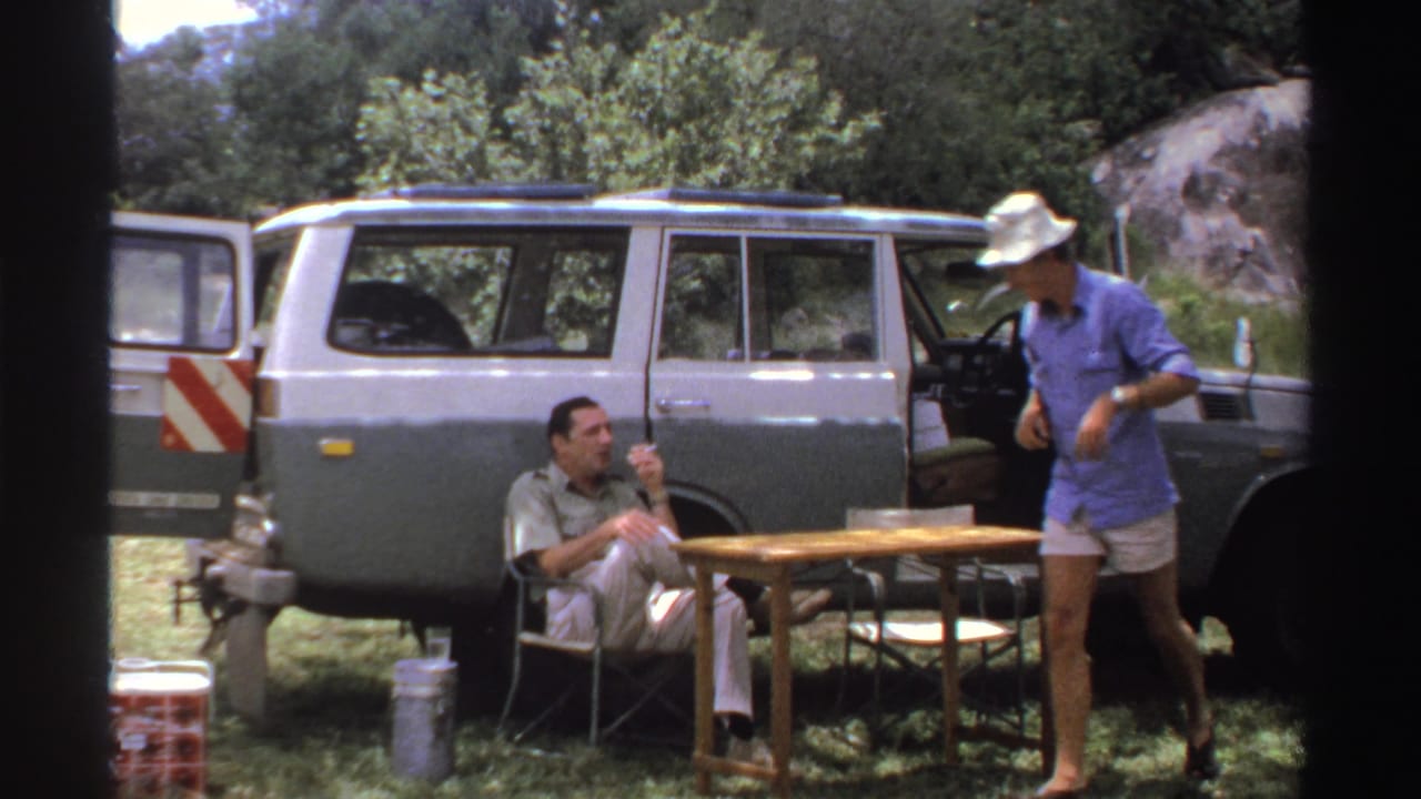 Two men beside an open vintage SUV, with one seated by a camp table and one standing in a hat.