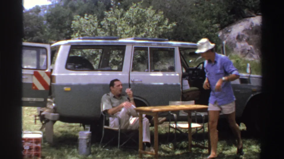 In 1969 Nigeria footage, two adult men prepare beside an open vintage SUV, with camp furniture and a cooler visible outdoors.
