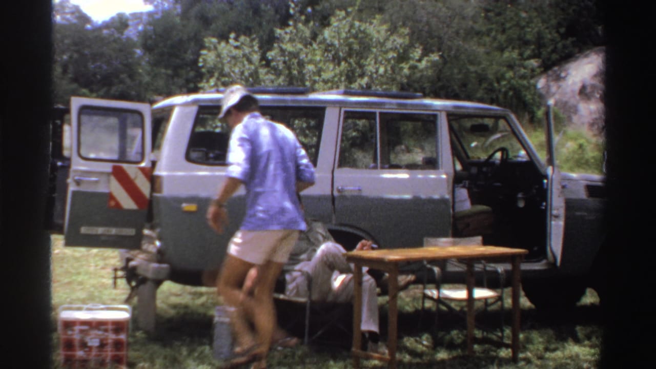 A man in a blue shirt and hat moves around a small camp setup beside an open vintage SUV in archival Nigeria travel footage.