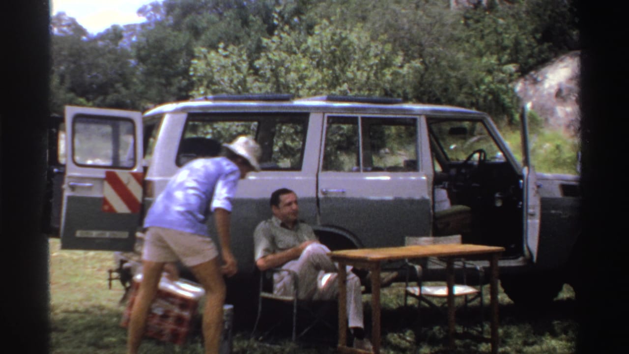 One adult man reaches near a red cooler while another sits in a folding chair beside the open SUV in 1969 Nigeria home-movie footage.