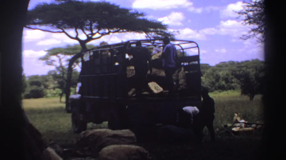 1969 Nigeria 8mm frame showing people beside a dark open-sided truck with sacks in a rural savanna setting.