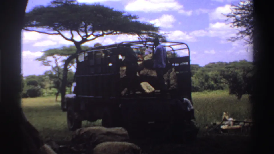 1969 Nigeria 8mm frame of a dark truck in savanna shade with people nearby and sacks lying in the foreground.