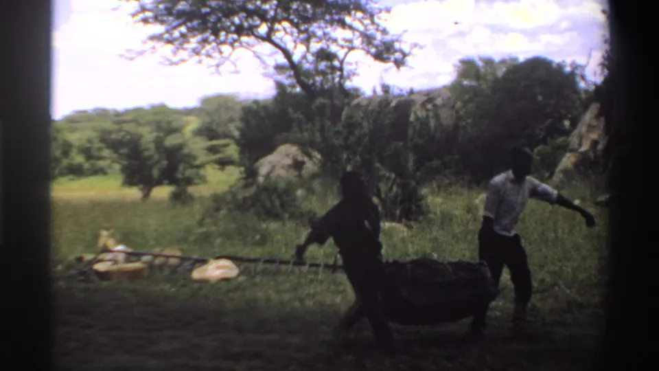 In Nigeria 1969 home-movie footage, two men move a heavy dark bundle through a grassy rural field beneath a bright sky.