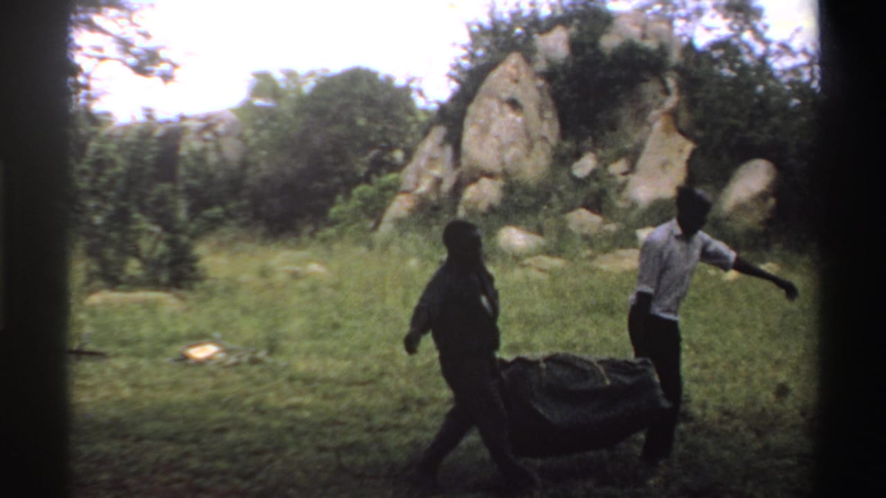Two men in a Nigeria 1969 8mm frame carry a bulky dark load across grass near trees and exposed rocks.