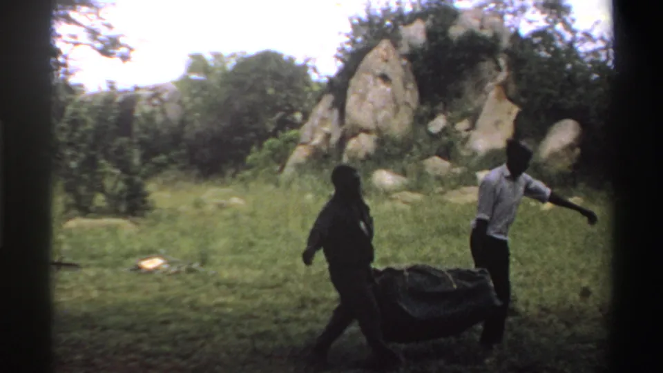 Two men in a Nigeria 1969 8mm frame carry a bulky dark load across grass near trees and exposed rocks.