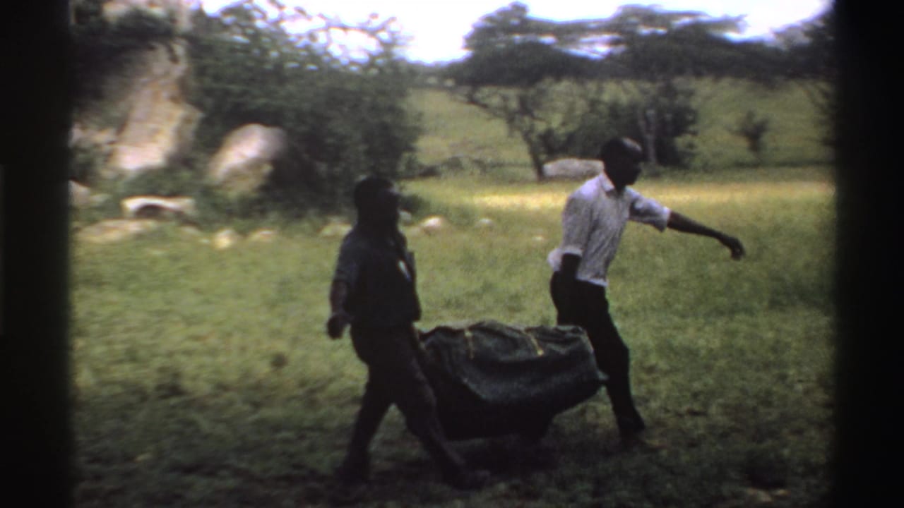 The handheld 8mm view shows two men carrying a heavy dark bundle through a grassy Nigeria landscape in 1969.