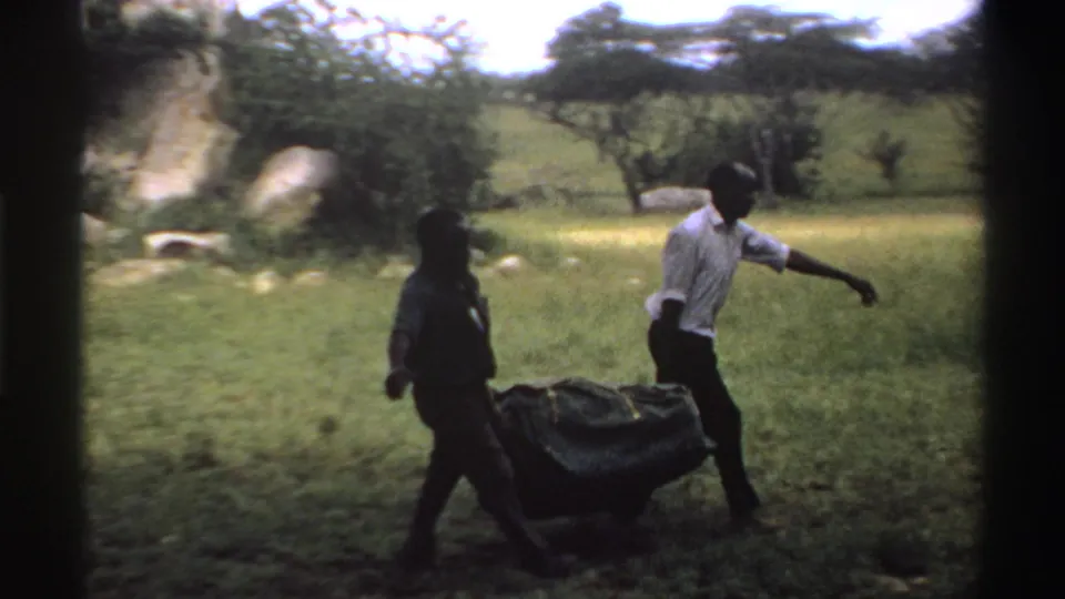 The handheld 8mm view shows two men carrying a heavy dark bundle through a grassy Nigeria landscape in 1969.