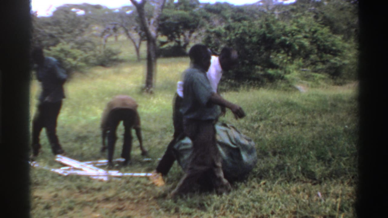 Men work together in the grass, with one bending near a low frame while others guide the green tarp.