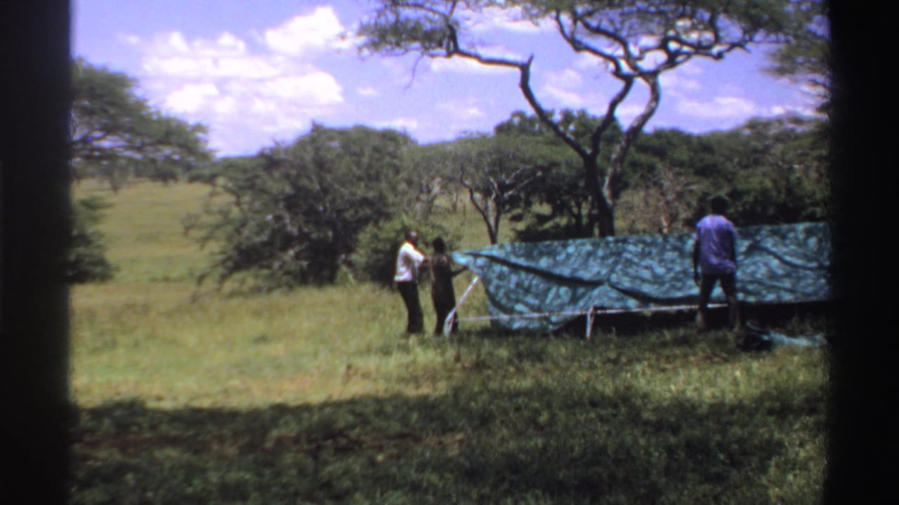 1969 Nigeria 8mm frame showing two adults at the side of a green tarp shelter with trees and open grassland behind them.