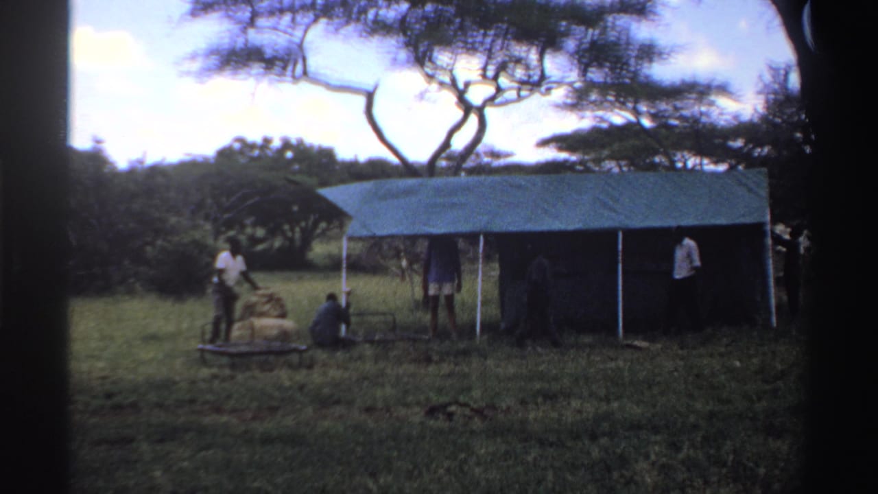 1969 Nigeria 8mm frame showing several men around a temporary blue shelter, with bundles and a metal frame in the grass.
