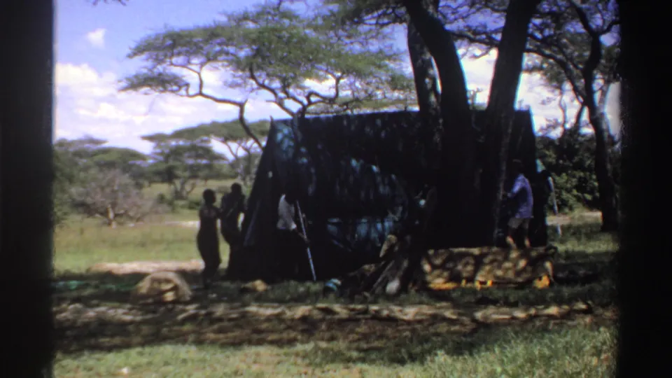 In Nigeria 1969 home-movie footage, several people gather near a dark tarp shelter beneath trees in a sunlit savanna landscape.