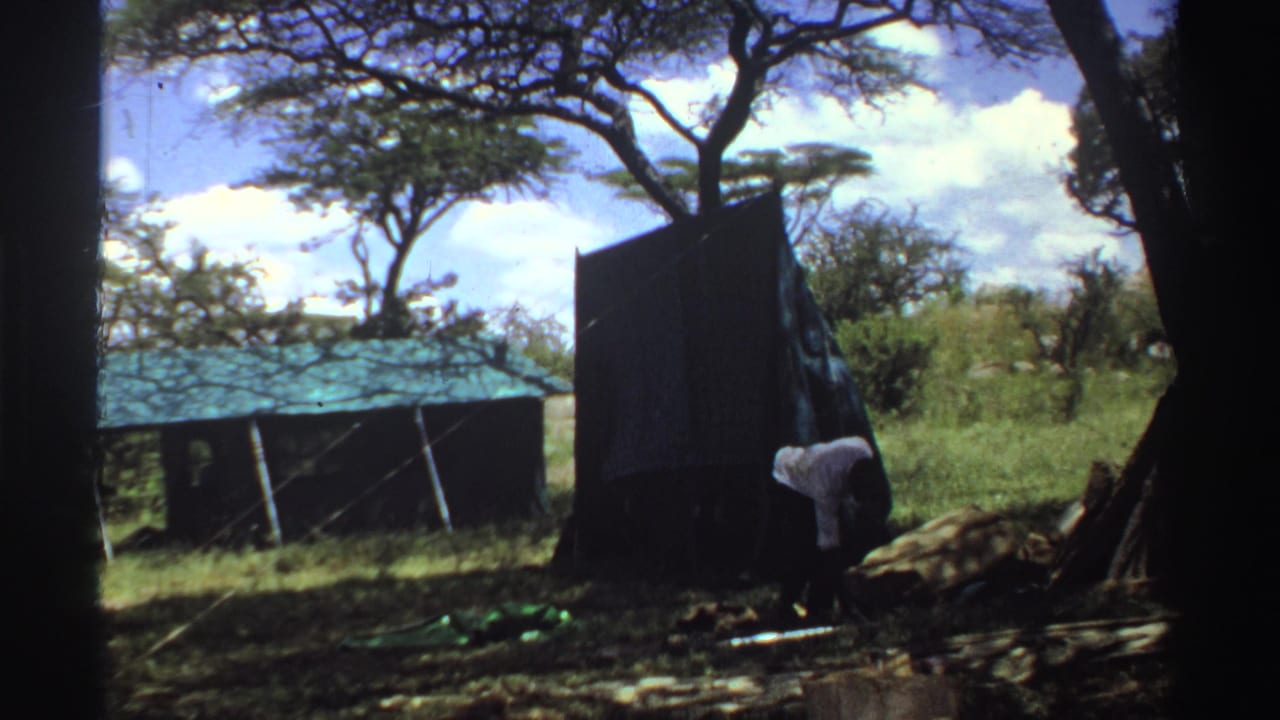 1969 Nigeria frame showing a person bent near a large upright tent or panel with trees and a roofed structure behind.