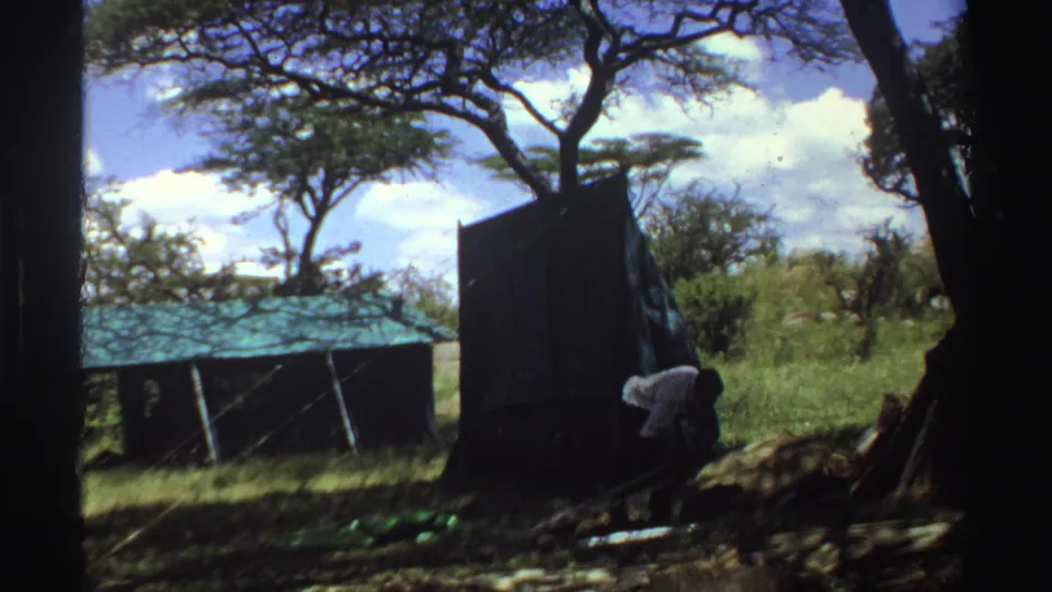 1969 Nigeria 8mm frame showing a dark tent, low green-roofed shelter, trees, and a person bending or moving near the tent edge.