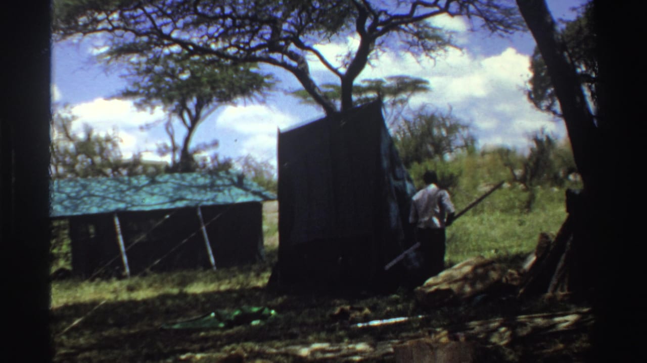 1969 Nigeria 8mm frame with a man in a light shirt carrying a long wooden pole or object toward figures near a dark tent.