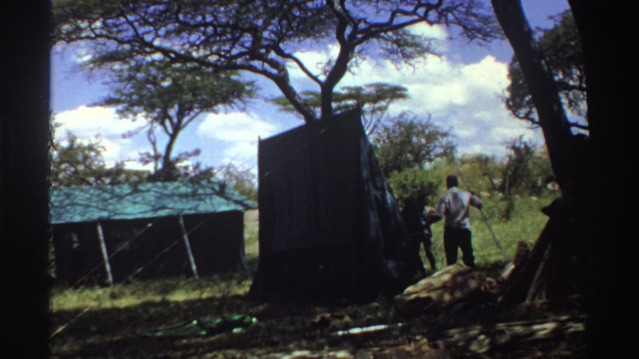 1969 Nigeria 8mm frame showing people gathered at a dark tent as a man holds a long pole or wooden piece in the grass.