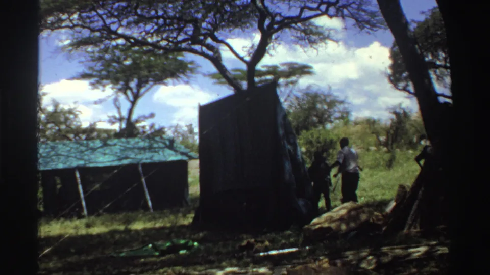 1969 Nigeria 8mm frame showing several people beside a dark tent in a grassy clearing, with trees and a low shelter behind them.